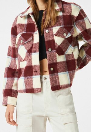Mujer con una chaqueta abotonada de cuadros rojos y blancos sobre un top corto negro y pantalones cargo blancos, de pie con una mano en la cadera.