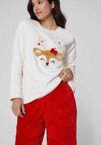 Biała bluza polarowa z nadrukiem twarzy renifera, ozdobiona haftowanymi detalami i czerwonymi akcentami, do kompletu czerwone pluszowe spodnie.