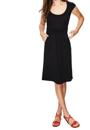 Femme portant une robe noire mi-longue à manches courtes et des sandales plates rouges à lanières, debout les mains dans les poches.