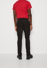 Černé cargo kalhoty s úzkým střihem, vybavené bočními kapsami, elastickým pasem a kontrastním prošíváním. K nim jsou černé boty.