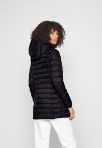 Svart boblejakke med hette, med en quiltet design og glatt tekstur. Lange ermer og en tettsittende silhuett. Hvite bukser synlige under.