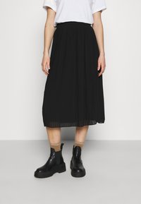 Sort plisseret midi-nederdel med elastisk talje, parret med sorte kraftige ankelstøvler og brune sokker. Båret med en hvid t-shirt.