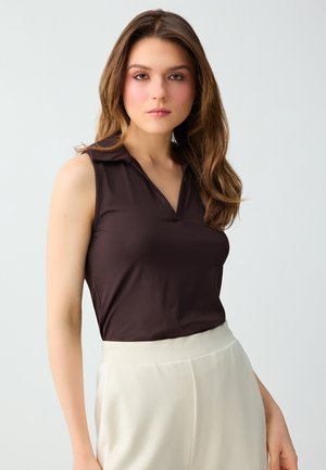 Giovane donna con lunghi capelli castani che indossa una canotta marrone scuro senza maniche e pantaloni bianchi a vita alta su uno sfondo chiaro e uniforme.