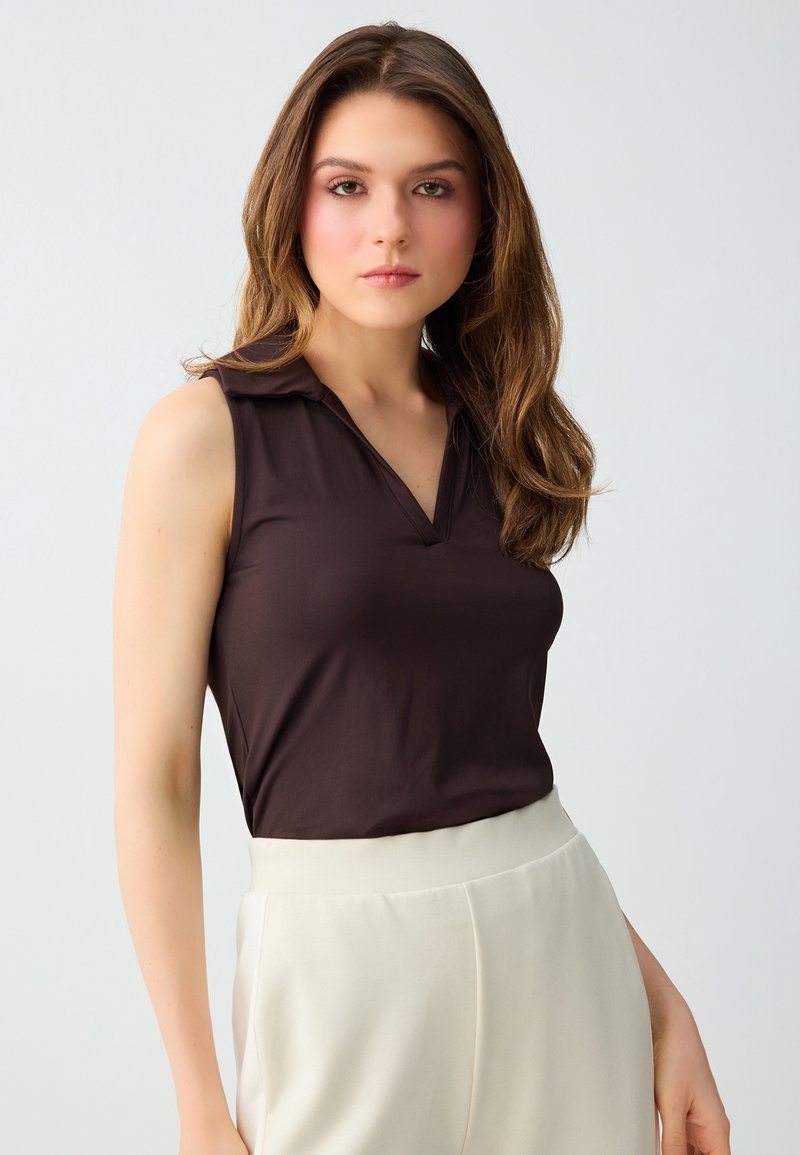 Jeune femme aux longs cheveux bruns portant un haut sans manches marron foncé et un pantalon blanc taille haute sur un fond clair uni.