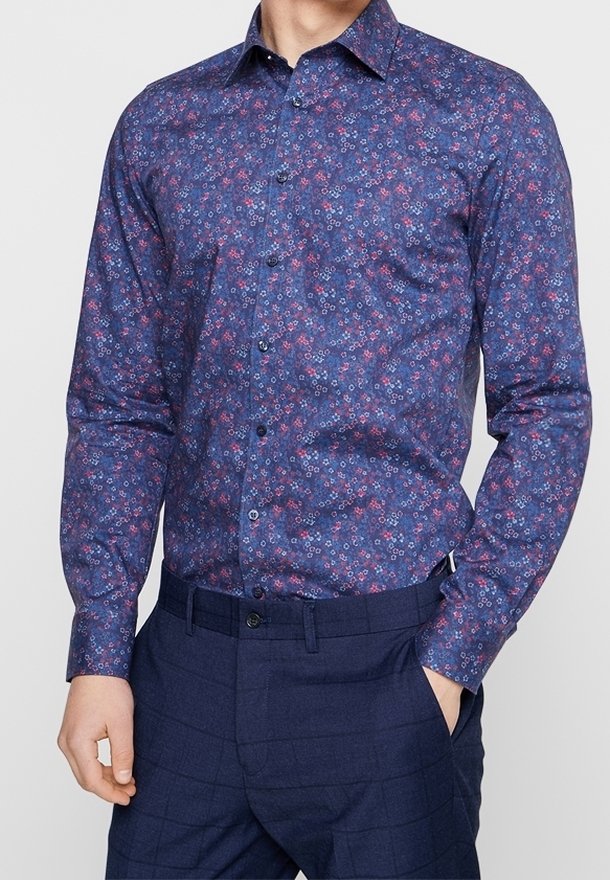 Homme portant une chemise bleu marine à manches longues avec un motif floral rouge et blanc, rentré dans un pantalon à carreaux bleu foncé.
