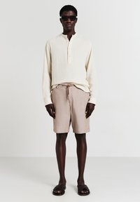 Hellbeige strukturierte Langarmbluse, braune gemusterte Shorts und schwarze Sandalen; Modell trägt dunkle Sonnenbrillen vor einem neutralen Hintergrund.
