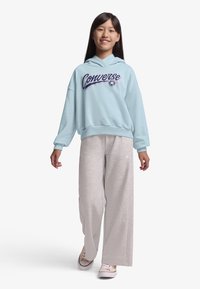 Φαρδιά γαλάζια φούτερ με κουκούλα με το λογότυπο "Converse" σε σκούρο μπλε, συνδυασμένη με γκρι φαρδιά φόρμα και ανοιχτόχρωμα αθλητικά παπούτσια. Λείο υφασμάτινο υλικό.