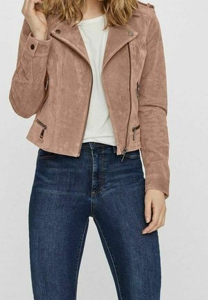 Kobieta w jasnobrązowej zamszowej kurtce motocyklowej na białej koszuli i ciemnoniebieskich jeansach z wysokim stanem, z ręką przy twarzy w luźnej pozie.