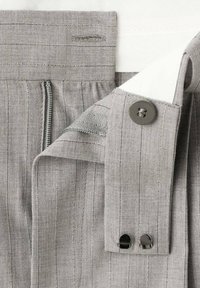 Pantalones de tela gris con finas rayas verticales, que presentan una cremallera, cierre de botón y detalles internos en la cinturilla. Textura suave.