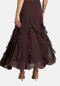 Gonna maxi in tessuto bordeaux con volanti a strati, caratterizzata da inserti trasparenti all'orlo. Indossata con sandali beige con cinturini.