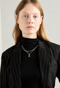 Gouden ketting met een smaragd hanger, gelaagd over een zwarte coltrui. De jas heeft een streepjespatroon in zwart en wit.