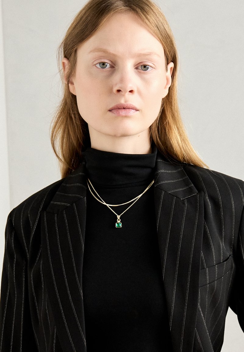 Gouden ketting met een smaragd hanger, gelaagd over een zwarte coltrui. De jas heeft een streepjespatroon in zwart en wit.