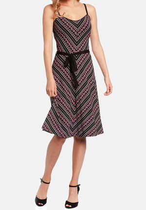 Woman wearing a sleeveless black dress with pink and white floral chevron pattern, tied black belt, and black open-toe heels.