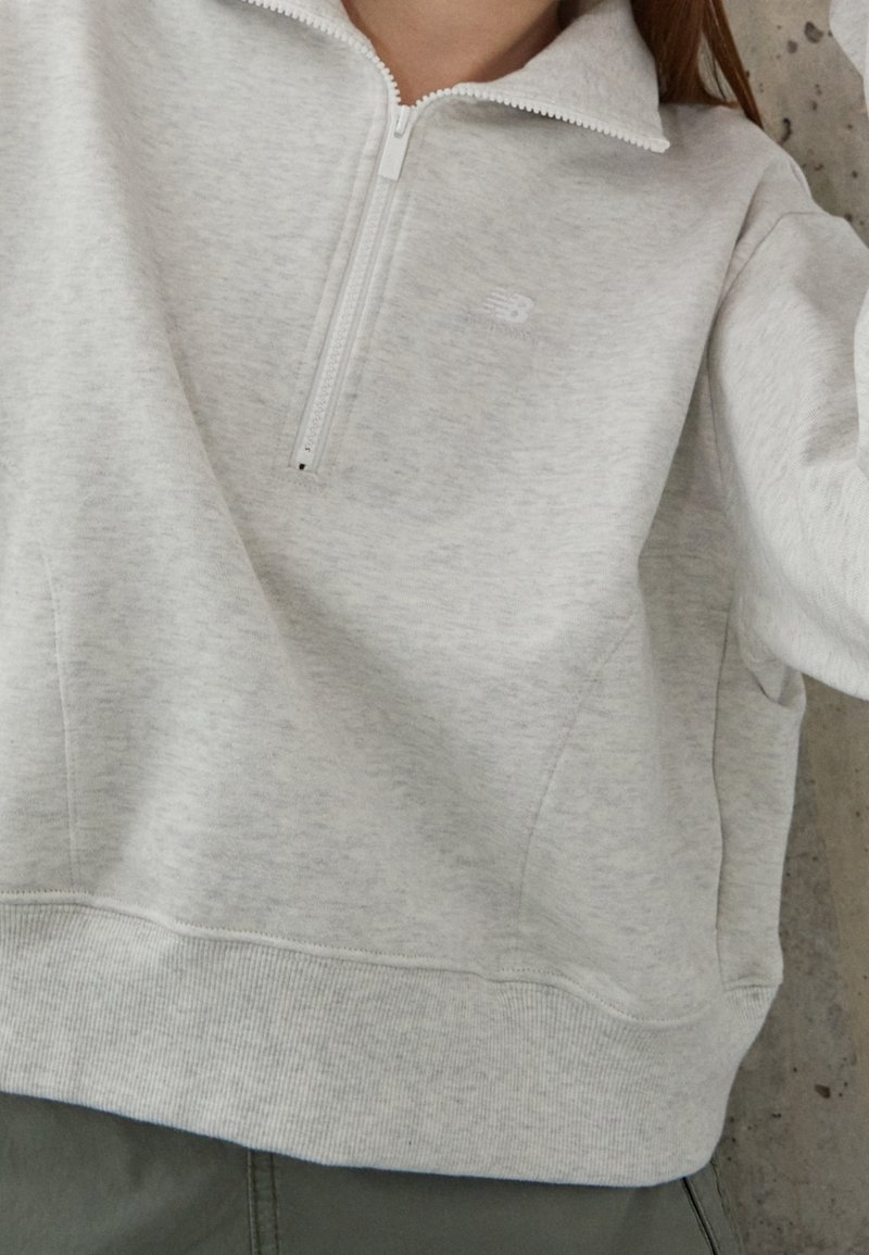 Personne portant un sweat-shirt gris clair à demi-fermeture éclair avec un petit logo sur la poitrine, associé à un pantalon foncé, sur un fond neutre.