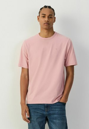 Junger Mann mit geflochtenem Haar, der ein schlichtes hellrosa T-Shirt und blaue Jeans trägt und mit neutralem Gesichtsausdruck vor einer weißen Wand steht.