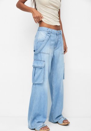 Person wearing light blue wide-leg denim cargo pants with side pockets and suspenders, paired with open-toe sandals and a beige cropped top.