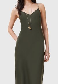 Olivgrön spaghetti axelbandsklänning med en figurnära byst och flödande kjol. Har en slät textur och en subtil lyster. Guldhalsband bärs.