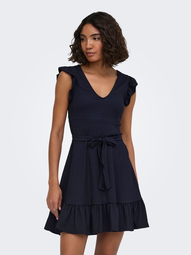 Femme aux cheveux courts et bouclés portant une robe noire sans manches avec des épaules volantées, un col en V, une ceinture nouée à la taille et un ourlet volanté.