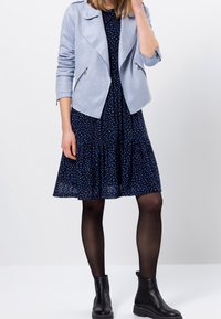 Veste en suede synthétique bleu clair avec zips argentés, portée sur une robe bleu marine à motif bleu dispersé, associée à des bottines noires.