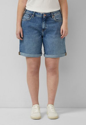Person trägt hochgekrempelte blaue Jeansshorts, weiße Sneaker und ein weißes Oberteil und steht vor einem einfarbigen grauen Hintergrund.