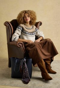 Strickpullover mit blauem und braunem Muster, braune Rock, beige Wildlederstiefel. Schwarze Tragetasche mit floralem Schal, sitzend in einem braunen Lederarmstuhl.