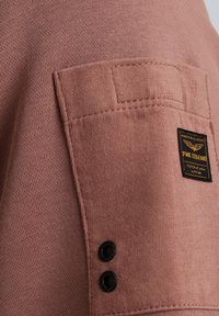 Camisa de algodón rosa de manga corta con un bolsillo de parche. Presenta botones negros y una etiqueta de tela en negro y dorado en el bolsillo izquierdo.
