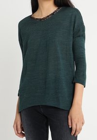 Dunkelblauer Strickpullover mit einem Bootsausschnitt und schwarzem Spitzenbesatz. Verfügt über Dreiviertelärmel und einen lockeren Schnitt mit abgerundetem Saum.