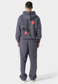 Přesný šedý oversized hoodie s kapucí na šňůrku, který má na zadní straně černý grafický design a červené akcenty; doplněný o odpovídající tepláky.