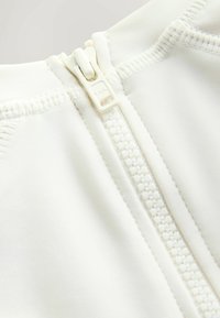 Detalle de una prenda blanca con cremallera, mostrando una tela lisa y texturizada con una cremallera de plástico en el centro, resaltando las costuras a lo largo de los bordes.