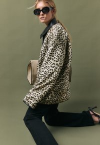Giacca in pelliccia sintetica con stampa leopardata e colletto in pelle nera, abbinata a pantaloni neri e sandali neri con tacco alto, mentre tiene una borsa beige.