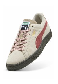 Beżowy zamszowy sneaker Puma z czerwoną paską, wzorzystymi sznurowadłami, czarną teksturowaną podeszwą oraz logo Puma na języku i boku.