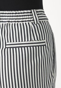 Schwarz-weißer, senkrecht gestreifter Stoff, mit einem flachen Bund vorne und einer Tasche. Das Material wirkt gewebt und strukturiert.
