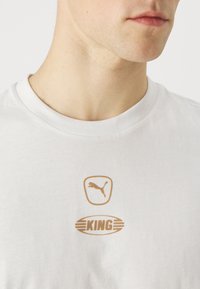 Witte katoen T-shirt met gouden bedrukte logo's: een springende poema in een vierkant en het woord "KING" in een horizontale ovaal. Korte mouwen en ronde hals.