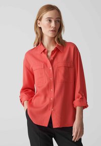 Camicia button-up color corallo con colletto, dotata di due tasche sul petto e maniche arrotolate. Realizzata in un tessuto morbido con una vestibilità comoda.