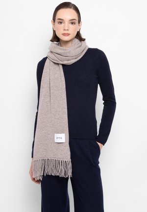 Sciarpa di lana morbida, beige chiaro con frange, drappeggiata intorno al collo. Etichette visibili sulla sciarpa. Modello indossa una maglia blu scuro.