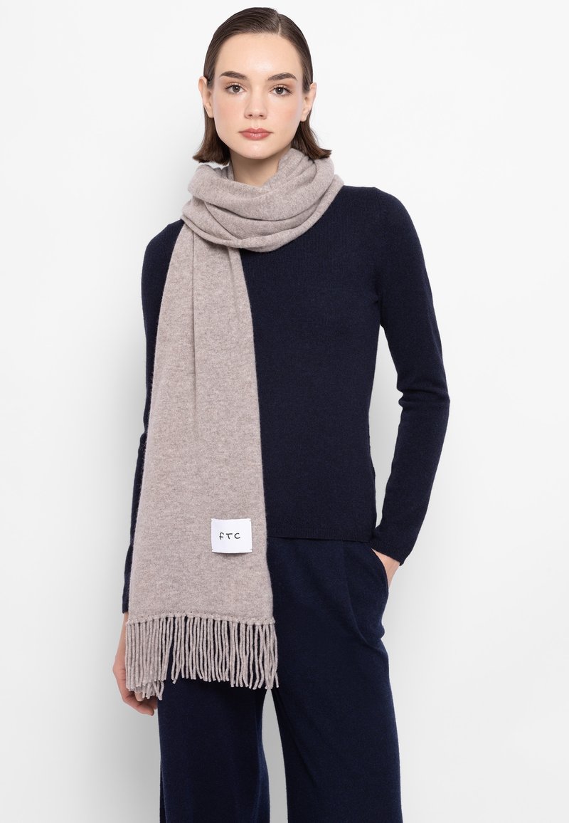 Miękki wełniany szal w jasnobeżowym kolorze z frędzlami, zarzucony na szyję. Na szalu widoczne metki. Modelka ma na sobie ciemnogranatową bluzkę.