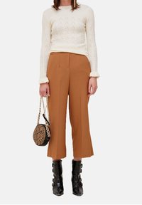 Kremowy sweter dzianinowy z teksturowanym wzorem, brązowe spodnie typu wide-leg i czarne botki za kostkę. Modelka trzyma okrągłą torbę z nadrukiem wężowym.