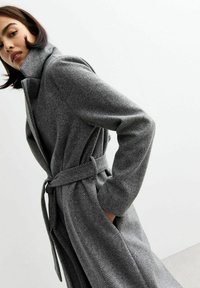 Manteau long à chevrons gris avec col et taille ceinturée, fabriqué dans un tissu texturé. Il présente des manches longues et une coupe décontractée.