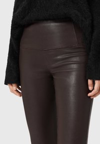 Brązowe legginsy z wysokim stanem wykonane z błyszczącego, przypominającego skórę materiału, z gładką fakturą i detalami w szwie w pobliżu pasa.