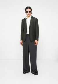 Olivgrüner, taillierter Blazer mit Revers, kombiniert mit weit geschnittenen grauen Hosen. Darunter ist ein weißes Hemd sichtbar; strukturierter Stoff und schwarze Schuhe.