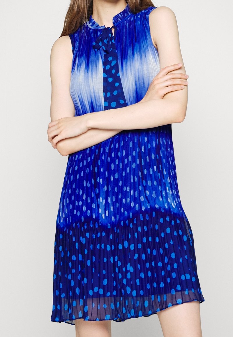 Femme portant une robe plissée bleue sans manches avec des motifs dégradés et à pois, bras croisés à la taille.