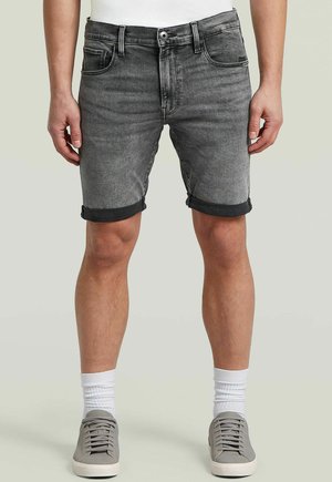 Mann trägt hochgekrempelte dunkelgraue Jeansshorts, weiße Socken und graue Turnschuhe, steht vor hellem Hintergrund.