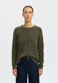 Pulover verde măsliniu, realizat dintr-un material moale și texturat, cu gât rotund și mâneci lungi, asortat cu blugi negri din denim și o curea neagră.