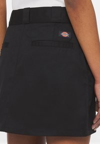Černá sukně Dickies s poutky na pásek a zadními kapsami, nošená s bílým topem na osobě se středně tmavou pletí.
