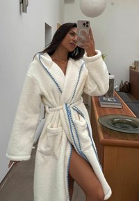 Balta plīša vanna halāts ar zilu apdari, garām piedurknēm, divām priekšējām kabatām un jostu. Audums izskatās mīksts un teksturēts.