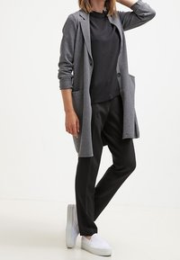 Šedý dlouhý cardigan s kapsami, vrstvený přes černý top, spárovaný s černými kalhotami a bílými nazouvacími botami. Hladká tkanina a přiléhavý střih.