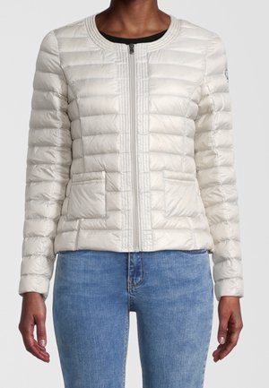 Femme portant une veste matelassée légère beige clair zippée avec deux poches avant et un jean bleu, visible du cou jusqu'aux hanches.