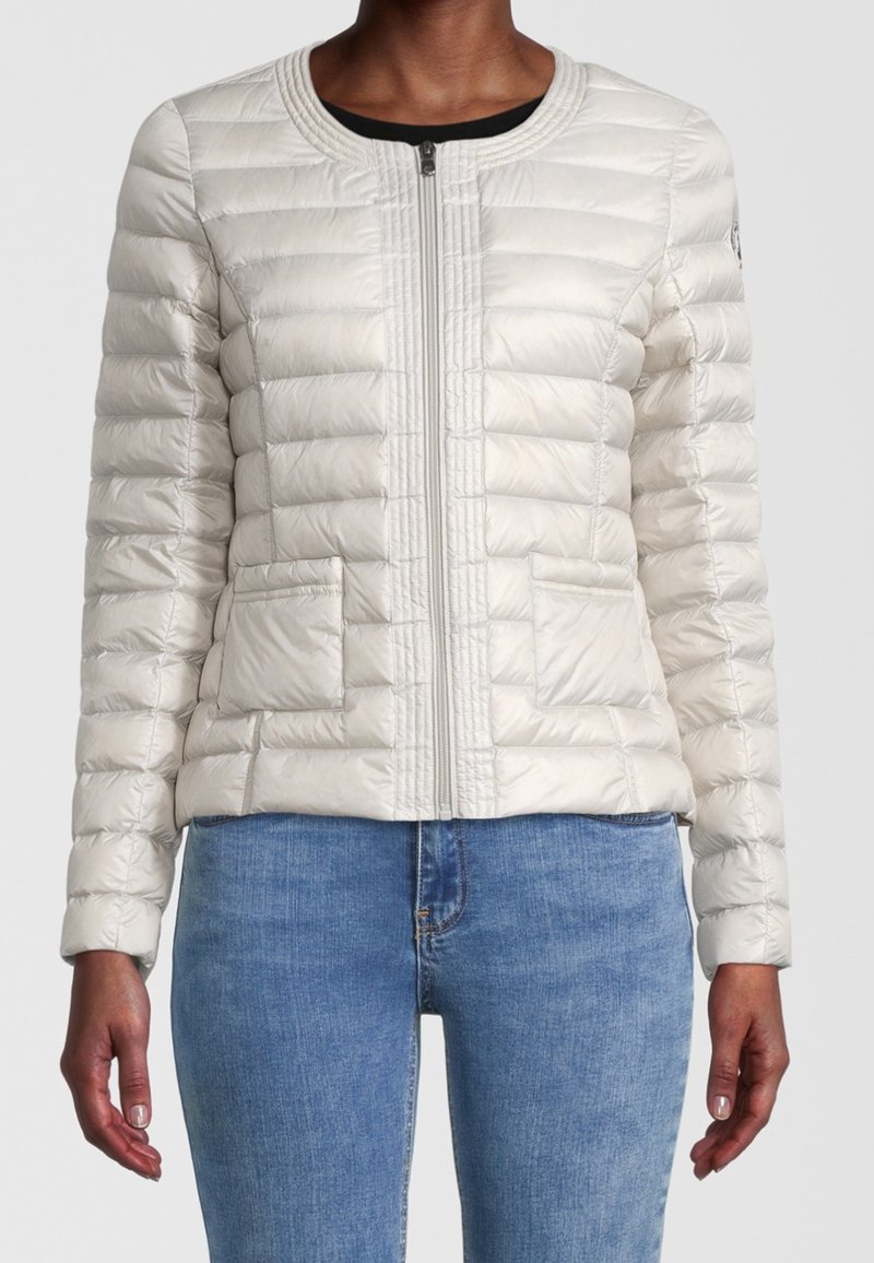 Femme portant une veste matelassée légère beige clair zippée avec deux poches avant et un jean bleu, visible du cou jusqu'aux hanches.