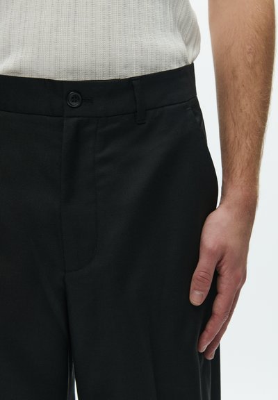 Sorte bukser med glat stof, en tilpasset talje med knaplukning og diskrete sidelommer. Tekstureret skjorte delvist synlig.