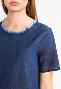Haut en denim avec un col rond et un col effiloché. Manches courtes, teinte bleu foncé, texture lisse et détail de couture subtil.
